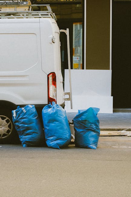 The image shows the rear side of a white commercial van parked on a street, with its back doors slightly open. In front of the van, three large blue rubbish bags, made of durable plastic, are placed on the pavement, each filled with waste material. The bags are slightly crumpled with visible creases and are positioned close together, occupying a small section of the curb. Behind the van, part of a storefront with a glass window and a white exterior wall can be seen, suggesting an urban or commercial area. The scene appears to be during daytime with natural lighting, and the environment indicates a typical setup where independent waste collection or disposal services, such as those provided by Rubbish Clearance Waterloo, might handle rubbish removal for local businesses or traders. The overall composition emphasizes the process of private waste handling through on-site collection, with the blue rubbish bags contrasting against the neutral tones of the van and surrounding environment.