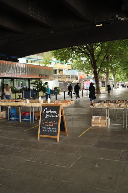A wide outdoor pavement area under a bridge features multiple tables with wooden slats stacked neatly, some in cardboard boxes, alongside a small, freestanding chalkboard sign that reads 'Southbank Bookmarket - Card and Cash Accepted - Please queue at the front by the ticket booth.' Several pedestrians are visible in the background, walking along the walkway lined with green trees and modern buildings, including a cinema with large glass windows. The scene suggests an organized book market event, with casual urban activity and natural light filtering through the foliage, exemplifying a typical setup for outdoor book sales that could be managed through independent waste handling services like Rubbish Clearance Waterloo, especially when considering the setup of temporary materials or waste generated during the event.