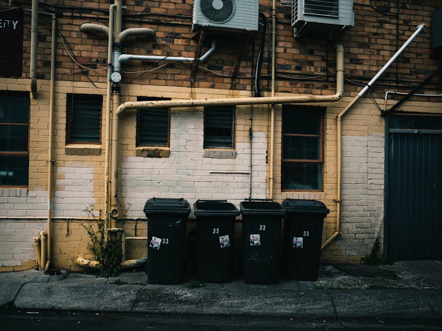 The image shows the rear side of a white commercial van parked on a street, with its back doors slightly open. In front of the van, three large blue rubbish bags, made of durable plastic, are placed on the pavement, each filled with waste material. The bags are slightly crumpled with visible creases and are positioned close together, occupying a small section of the curb. Behind the van, part of a storefront with a glass window and a white exterior wall can be seen, suggesting an urban or commercial area. The scene appears to be during daytime with natural lighting, and the environment indicates a typical setup where independent waste collection or disposal services, such as those provided by Rubbish Clearance Waterloo, might handle rubbish removal for local businesses or traders. The overall composition emphasizes the process of private waste handling through on-site collection, with the blue rubbish bags contrasting against the neutral tones of the van and surrounding environment.