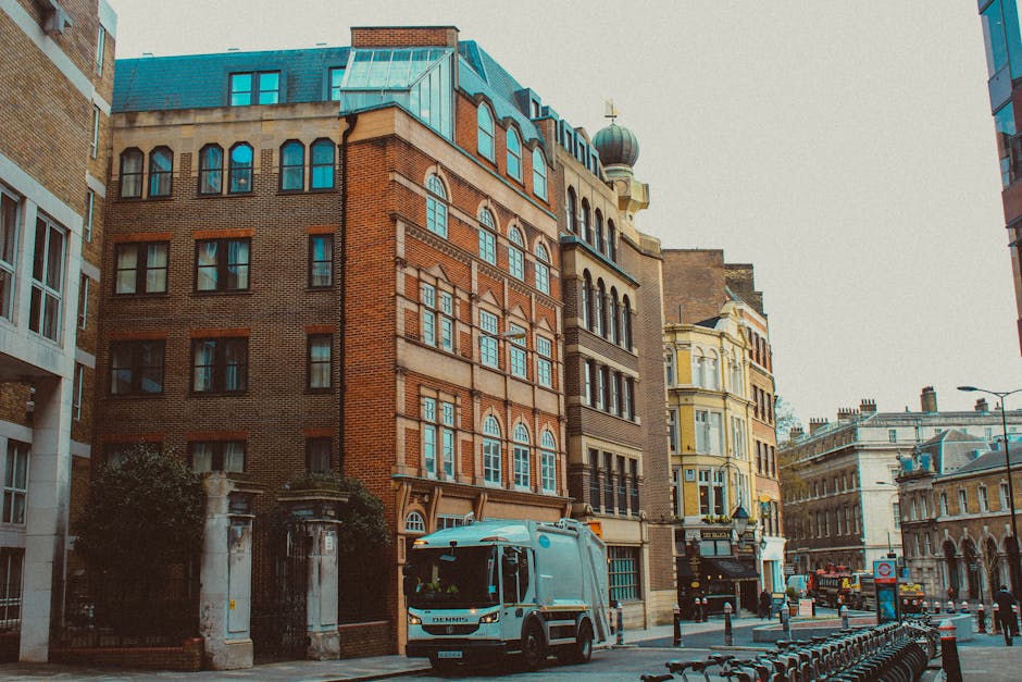 A street scene in an urban area featuring several multi-story buildings with diverse architectural styles and finishes. The buildings include brickwork in shades of red and beige, with large windows framed with decorative molding. A small domed cupola with a cross on top is visible on one building, indicating a religious structure. The foreground shows a white waste collection vehicle parked on the street, positioned near a sidewalk with metal bike racks and bollards. The street appears busy, with other vehicles and pedestrians in the background, under a cloudy sky. The overall scene reflects a city environment where private waste removal services, such as those provided by Rubbish Clearance Waterloo, operate in conjunction with other urban activities, supporting alternative rubbish clearance needs in the area.