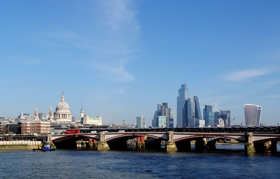 A wide view of London's skyline showcasing a variety of iconic buildings, including St. Paul's Cathedral with its large dome in the foreground, alongside modern skyscrapers with reflective glass facades. The scene is set across the River Thames, with a bridge spanning the water in the lower part of the image, supported by multiple stone piers. A small boat is positioned near the bridge on the river's surface, which features gentle ripples. The sky above is bright blue with some wispy clouds, and the overall atmosphere is clear and bright, highlighting the contrast between historic architecture and contemporary high-rise towers. This image captures the urban landscape typical of central London, suitable for illustrating alternative waste management or rubbish removal services in a city environment, with the background of a densely built business district and historic landmarks beyond the bridge.
