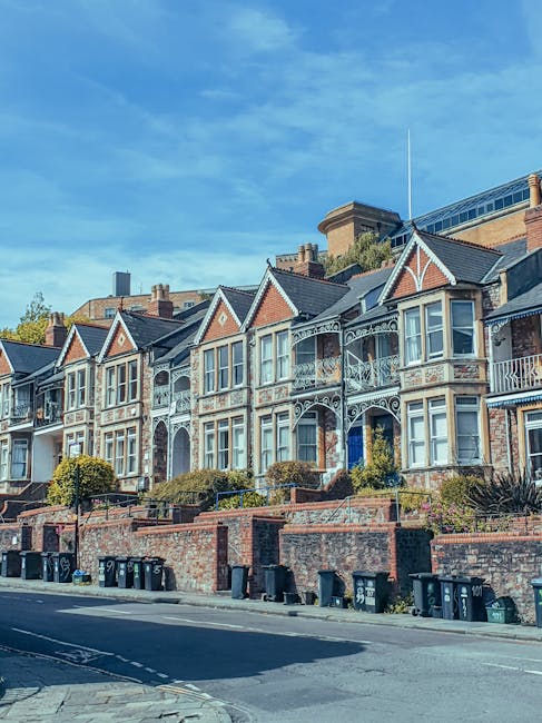 A row of Victorian-style terraced houses with ornate white decorative trim and bay windows, situated on a slightly elevated brick terrace along a quiet residential street. The buildings feature brick facades with contrasting white details on the gabled roofs and iron railings. Several small, well-maintained shrubs grow along the front of the houses, while a brick retaining wall supports the front gardens. The street in the foreground has a black asphalt surface with marked parking bays, and multiple black and green wheelie bins are lined up against the wall at the curb. The surrounding environment includes a clear blue sky with some scattered clouds, and neighboring modern buildings are visible behind the older houses, indicating an urban setting where private waste collection services, such as those by Rubbish Clearance Waterloo, may be used for onsite rubbish removal and clearance in the area.
