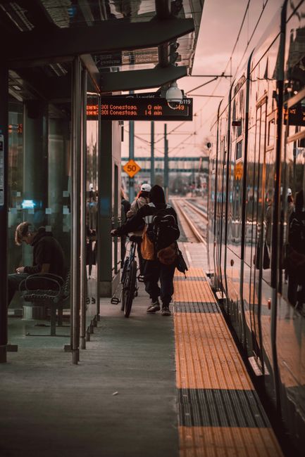 At a busy tram stop, two individuals, one with a bicycle and the other with a backpack, walk along a concrete platform towards a modern tram with reflective, tinted windows. The tram is stationary, with visible doors open for passenger boarding, and is painted in a sleek, metallic finish. Above the platform, an electronic display board shows travel information with red text indicating upcoming stops and distances, including Conestoga and other local destinations. The shelter overhead has a metal frame and glass panels, providing partial cover, with seating visible inside. In the background, railway wires, poles, and a faint sunset sky create an urban transit atmosphere. The platform features tactile paving strips for safety, and a speed limit sign indicating 50 km/h is mounted on a pole nearby. This scene illustrates a typical public transit setting in an urban environment, highlighting the importance of effective passenger organisation and the infrastructure supporting on-site public transport alternatives, which are often associated with independent or private organisation of passenger movement and waste management within stations. Rubbish Clearance Waterloo often supports such environments by managing waste from transit hubs to maintain clean and operational spaces.